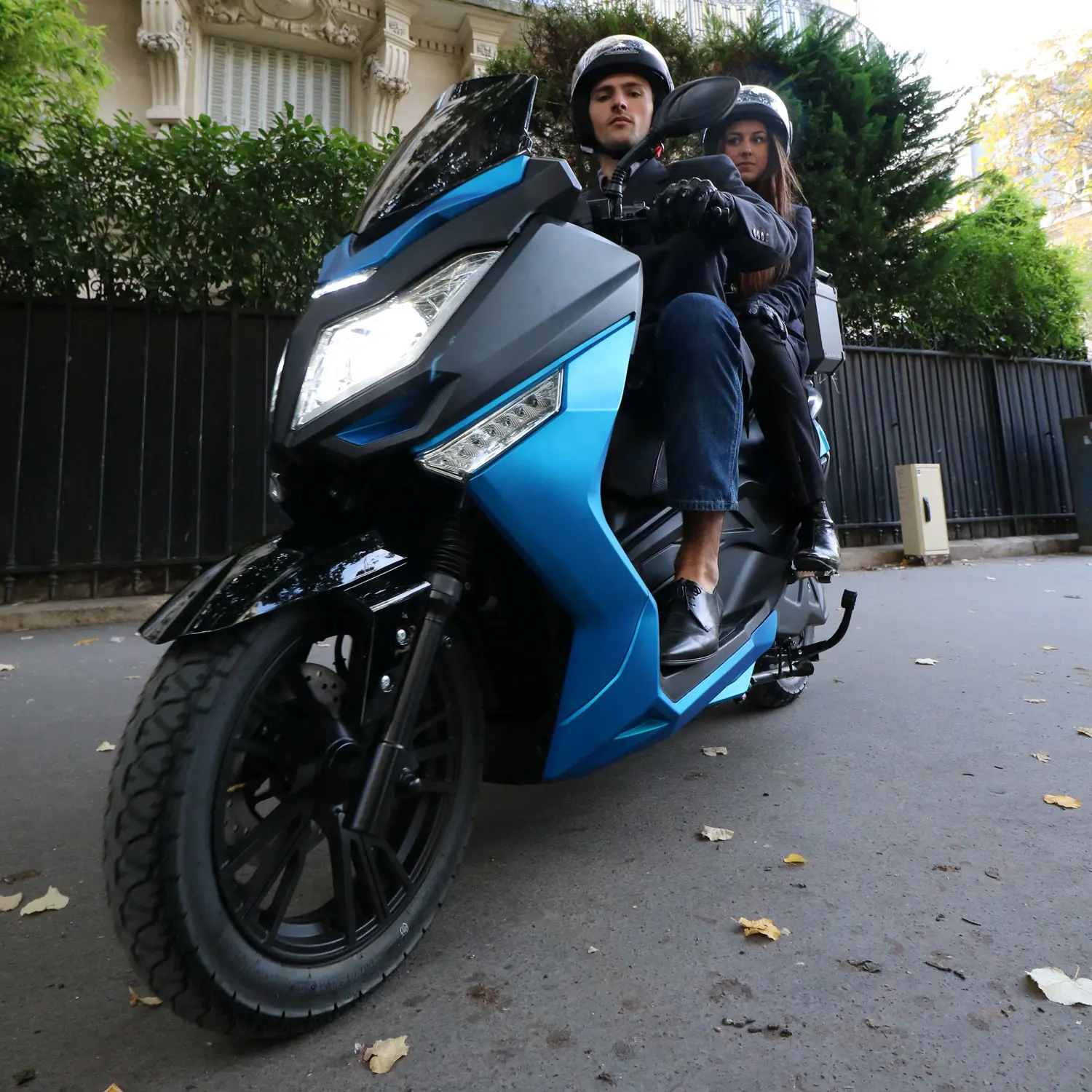 Scooter électrique Pink Mobility Pink Fly Phase 3 bleu en circulation avec deux passagers, en milieu urbain.